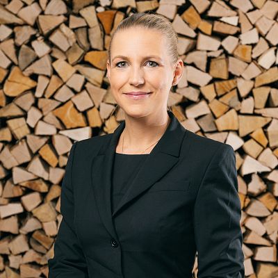 Frau in schwarzem Blazer vor gestapeltem Brennholz. Sie hat blondes Haar und lächelt in die Kamera. Der Hintergrund zeigt viele Holzscheite in gleichmäßiger Anordnung.