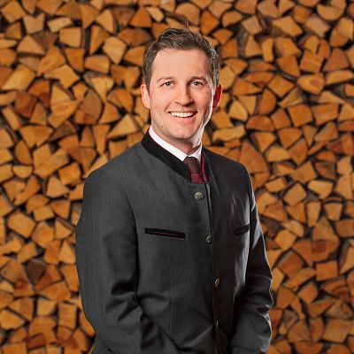 Ein Mann in traditioneller Kleidung lächelt vor einem Hintergrund aus gestapeltem Kaminholz. Er trägt einen Anzug mit einer dunklen Jacke und einem Hemd.