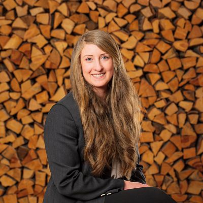 Eine Frau mit langem Haar sitzt in einem schwarzen Blazer vor einem gestapelten Holzscheitelhintergrund und lächelt freundlich in die Kamera.