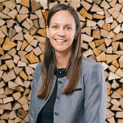 Frau mit langen braunen Haaren lächelt vor einem gestapelten Holzstapel. Sie trägt eine graue Jacke und ein schwarzes Oberteil. Der Hintergrund wirkt rustikal.