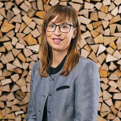 Eine Person mit Brille und braunem Haar lächelt, während sie vor einem Hintergrund steht, der aus gestapeltem Brennholz besteht. Sie trägt eine graue Jacke.
