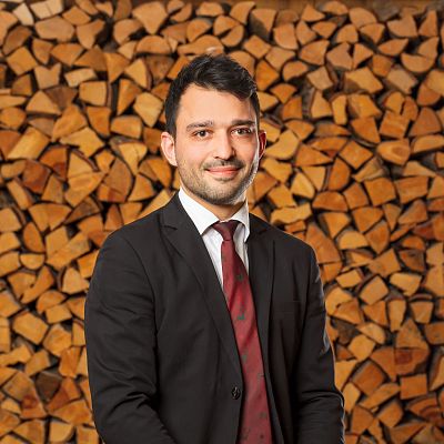 Ein Mann in einem schwarzen Anzug und roter Krawatte steht vor einem Stapel aufgeschichteter Holzscheite. Er hat dunkles Haar und lächelt leicht.