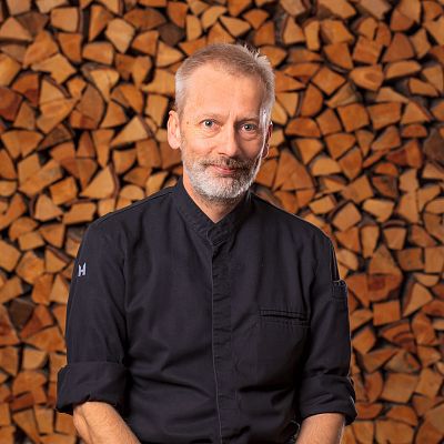 Ein Mann mit weißem Haar und Bart steht vor einer Wand aus ordentlich gestapeltem Kaminholz. Er trägt eine schwarze Kochjacke und blickt ernst in die Kamera.