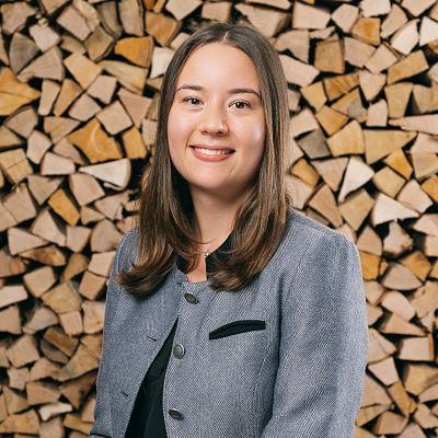 Eine lächelnde Person in einem grauen Blazer steht vor einem Stapel gestapelter Holzscheite. Der Hintergrund besteht aus ordentlich gestapeltem Feuerholz.