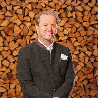 Ein lächelnder Mann in traditioneller bayerischer Kleidung steht vor einer großen Menge gestapeltem Holz. Er trägt ein dunkles Sakko und ein Einstecktuch.