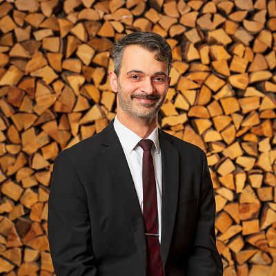 Ein Mann in einem schwarzen Anzug mit roter Krawatte steht vor einem Hintergrund aus gestapeltem Brennholz. Er lächelt sanft und hat glattes, graues Haar.