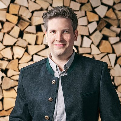 Ein Mann in traditioneller Tracht mit dunkler Jacke steht lächelnd vor einem gestapelten Holzstapel. Er hat kurzes Haar und trägt ein helles Hemd.