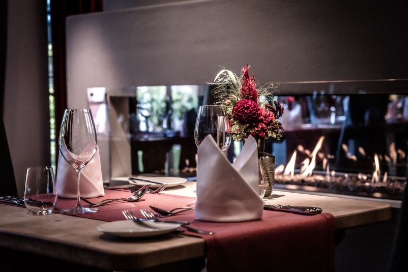 Stilvoll gedeckter Tisch in einem Restaurant mit gefalteten weißen Servietten, Wein- und Wassergläsern, Besteck und einem Blumenarrangement. Im Hintergrund ein Kamin.