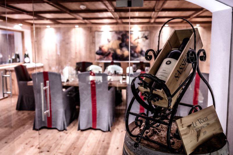 Elegantes Esszimmer mit Holzwänden und Holzboden, dekoriert mit einem Weinregal im Vordergrund. Stühle sind mit grauen Überzügen und roten Schleifen versehen.
