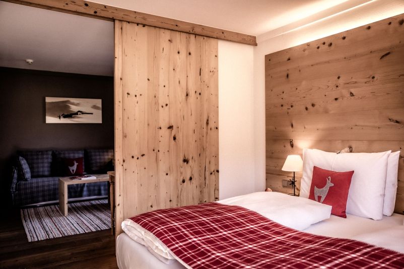 Gemütliches Hotelzimmer mit einem großen Bett, kariertem Überwurf und passendem Kissen. Eine Schiebetür öffnet den Blick auf einen Wohnbereich.