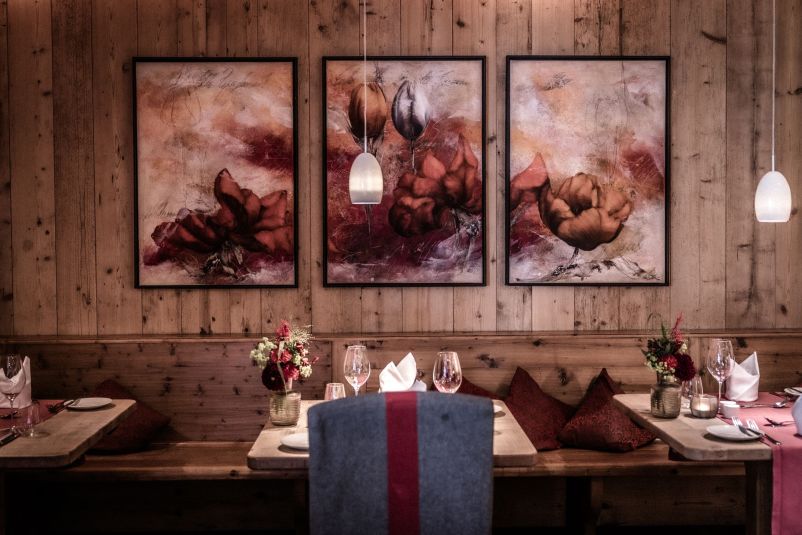 Gemütliches Restaurant mit Holzwänden, elegantem Tischgedeck und romantischer Beleuchtung. Drei große Blumengemälde schmücken die Wand über den Tischen.