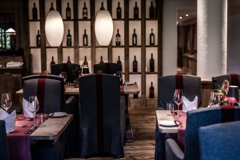 Elegantes Restaurant mit Holzelementen, stilvoll gedeckten Tischen und stimmungsvoller Beleuchtung. Im Hintergrund eine Wand mit dekorativen Weinflaschen.