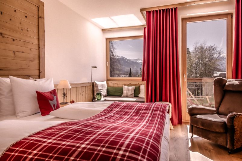 Gemütliches Hotelzimmer mit Holzverkleidung, rotem Karo-Deckenüberwurf und großen Fenstern mit Blick auf die Berge. Ein brauner Sessel steht in der Ecke.
