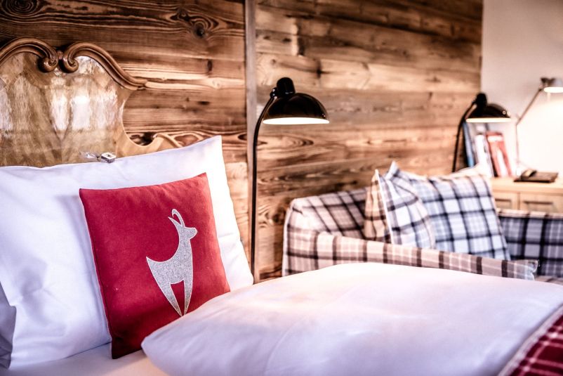 Gemütliches Schlafzimmer mit Holzverkleidung, bequemen Bett mit weißen Kissen und rotem Kissen mit Hirschmotiv. Neben dem Bett steht eine karierte Couch.