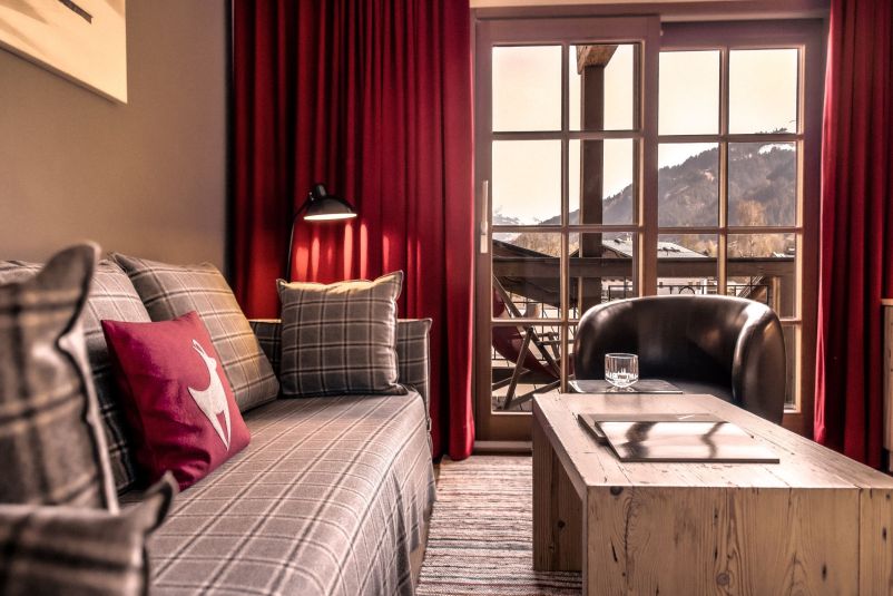 Gemütliches Wohnzimmer mit einem Sofa in Karo-Muster, roten Vorhängen, einem Ledersessel und einem Holztisch. Durch Fenster sieht man Berge in der Ferne.