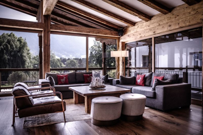 Gemütlicher Wohnraum mit Holzbalkendecke, großen Fenstern und moderner Einrichtung. Dunkle Sofas, ein Holztisch und helle Hocker sorgen für harmonischen Kontrast.