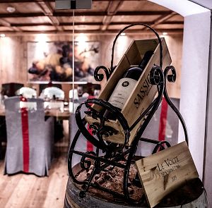 Elegantes Esszimmer mit Holzwänden und Holzboden, dekoriert mit einem Weinregal im Vordergrund. Stühle sind mit grauen Überzügen und roten Schleifen versehen.