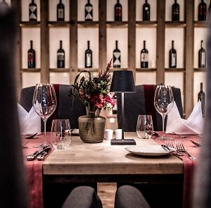 Elegantes Restaurant mit einem Holzregal voller Weinflaschen im Hintergrund. Der Tisch ist mit einem Blumenarrangement, Weingläsern und Besteck dekoriert.