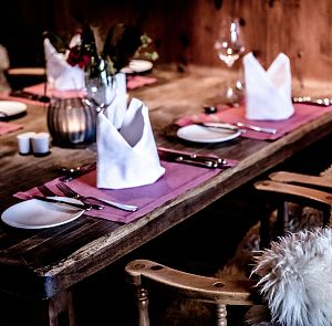 Gemütlicher rustikaler Esstisch mit flauschigen Fellstühlen, lila Platzsets, weiß gefalteten Servietten und elegantem Geschirr. Dezente Beleuchtung.