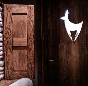 Ein rustikaler Raum mit gestapelten weißen Handtüchern in einem Holzregal. Rechts sieht man eine Tür mit einem Hirschsymbol, das in das Holz eingelassen ist.
