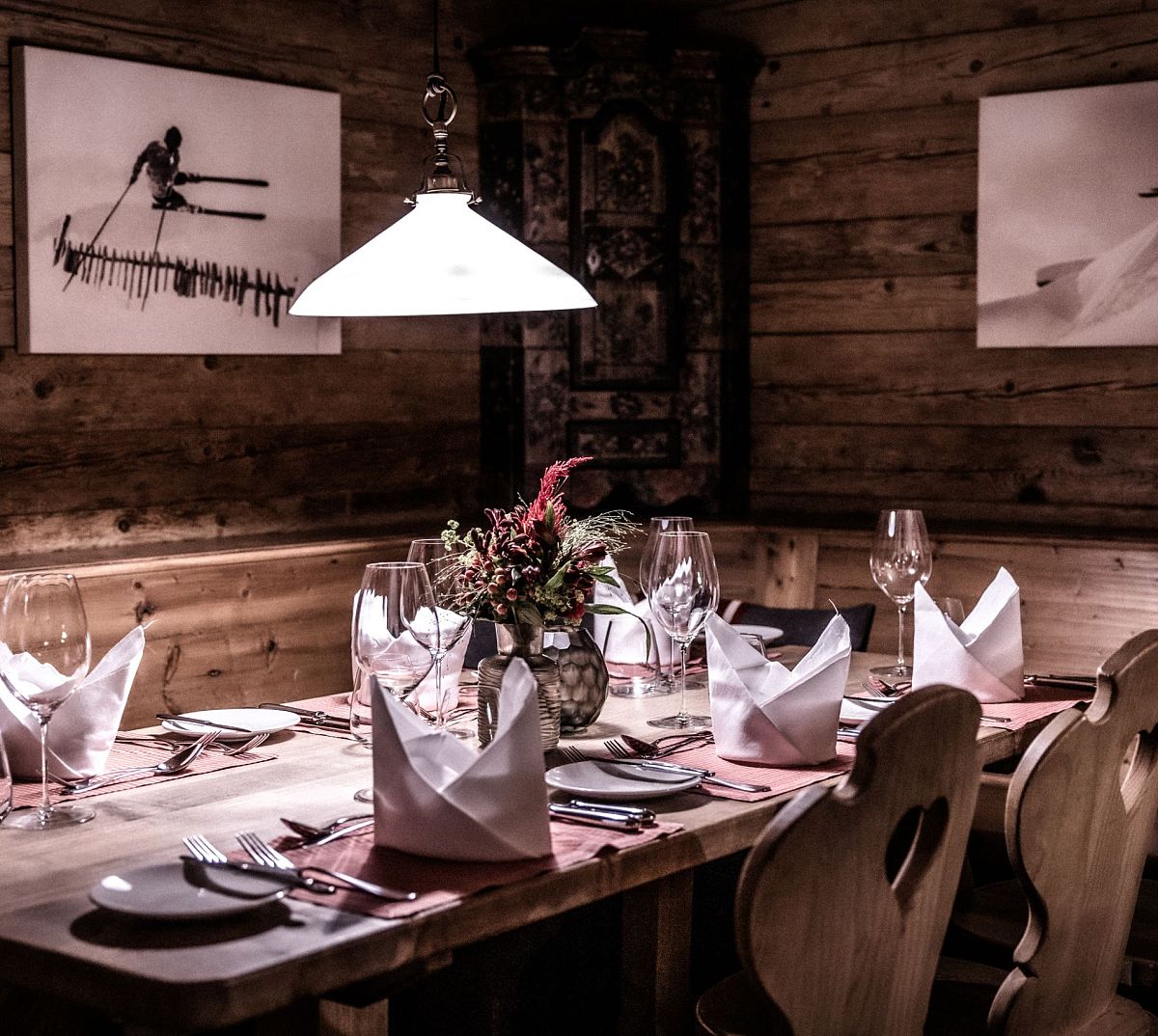 Gemütliches Restaurant mit Holzwänden, dekoriert mit Ski-Bildern. Ein gedeckter Tisch mit Glaswaren, Servietten und Blumengesteck unter einer Hängelampe.