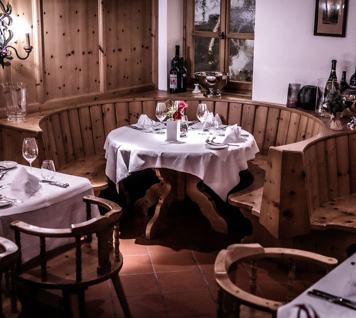 Gemütliches Restaurant mit Holzmöbeln, runden Tischen und weißen Tischdecken. Weinflaschen stehen auf den Tischen und Regalen als Dekoration.