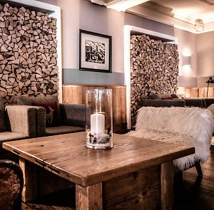 Gemütlicher Raum mit Holzwänden und gestapeltem Kaminholz. Gemütliche Sofas mit Kissen, Holztisch mit Kerze in Glas. Warmes, einladendes Ambiente.
