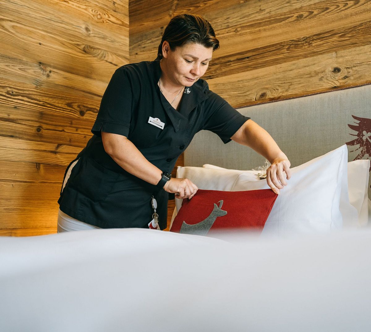 Eine Frau richtet ein Kissen in einem Hotelzimmer her. Sie trägt eine schwarze Uniform. Die Holzwände und das rote Kissen mit Adler-Motiv wirken gemütlich.