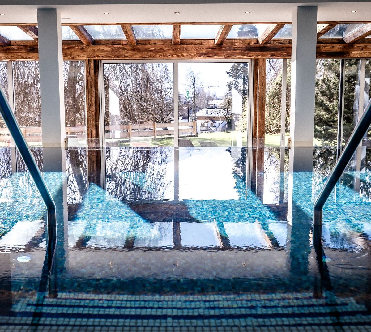 Innenpool mit glitzerndem Wasser, großen Fensterfronten und Blick auf einen Wintergarten mit kahlen Bäumen. Holzbalken und Tageslicht schaffen eine entspannte Atmosphäre.