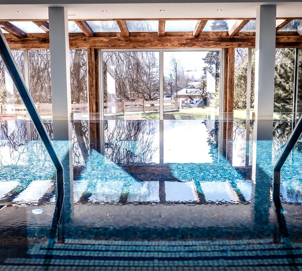 Innenpool mit glitzerndem Wasser, großen Fensterfronten und Blick auf einen Wintergarten mit kahlen Bäumen. Holzbalken und Tageslicht schaffen eine entspannte Atmosphäre.