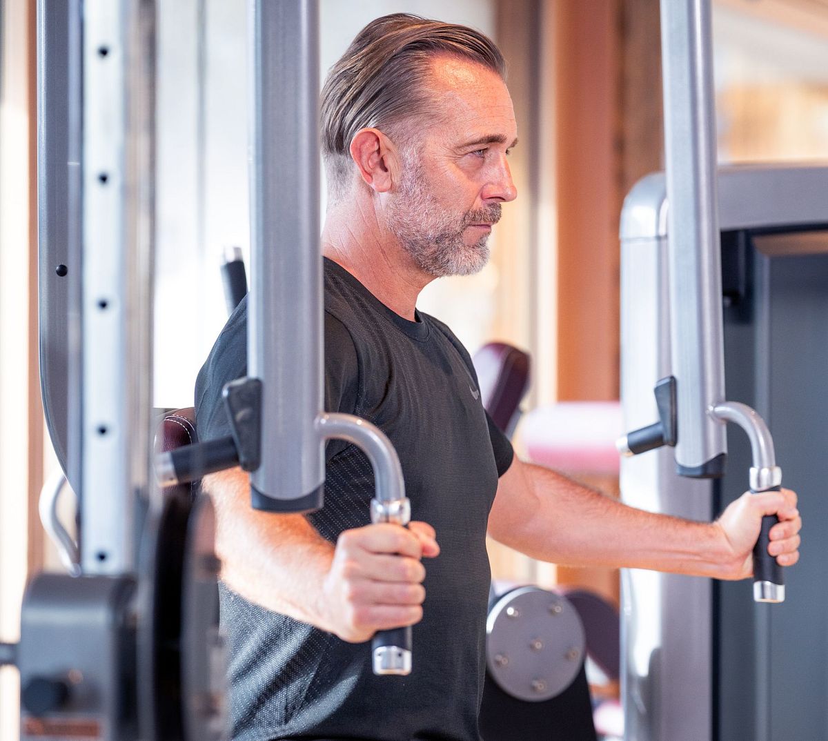 Ein Mann mittleren Alters trainiert an einer Brustpresse im Fitnessstudio. Er trägt ein schwarzes T-Shirt und konzentriert sich auf seine Übung.