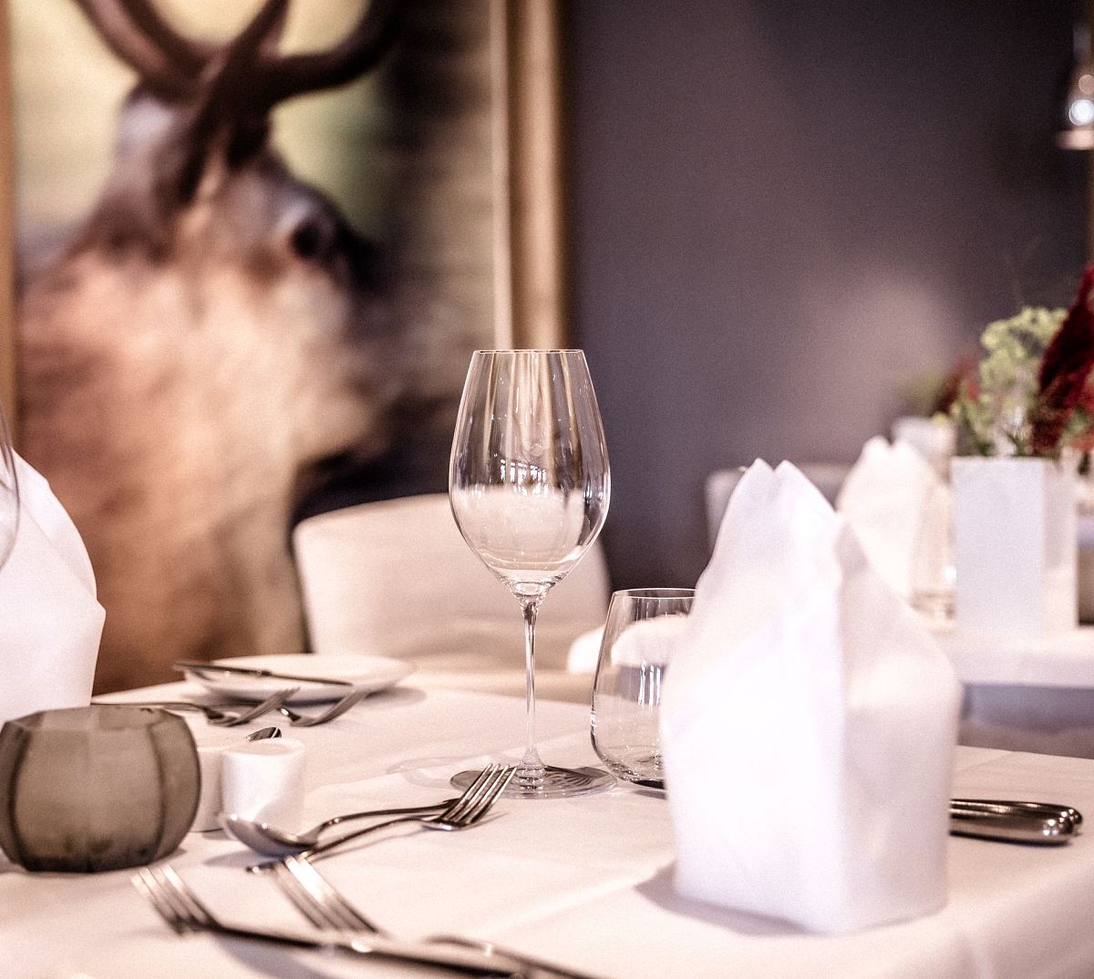Elegantes Restaurant mit weißen Tischdecken, gestärkten Servietten und edlem Besteck. Im Hintergrund ein großes Hirschbild. Dezente Blumendekorationen.