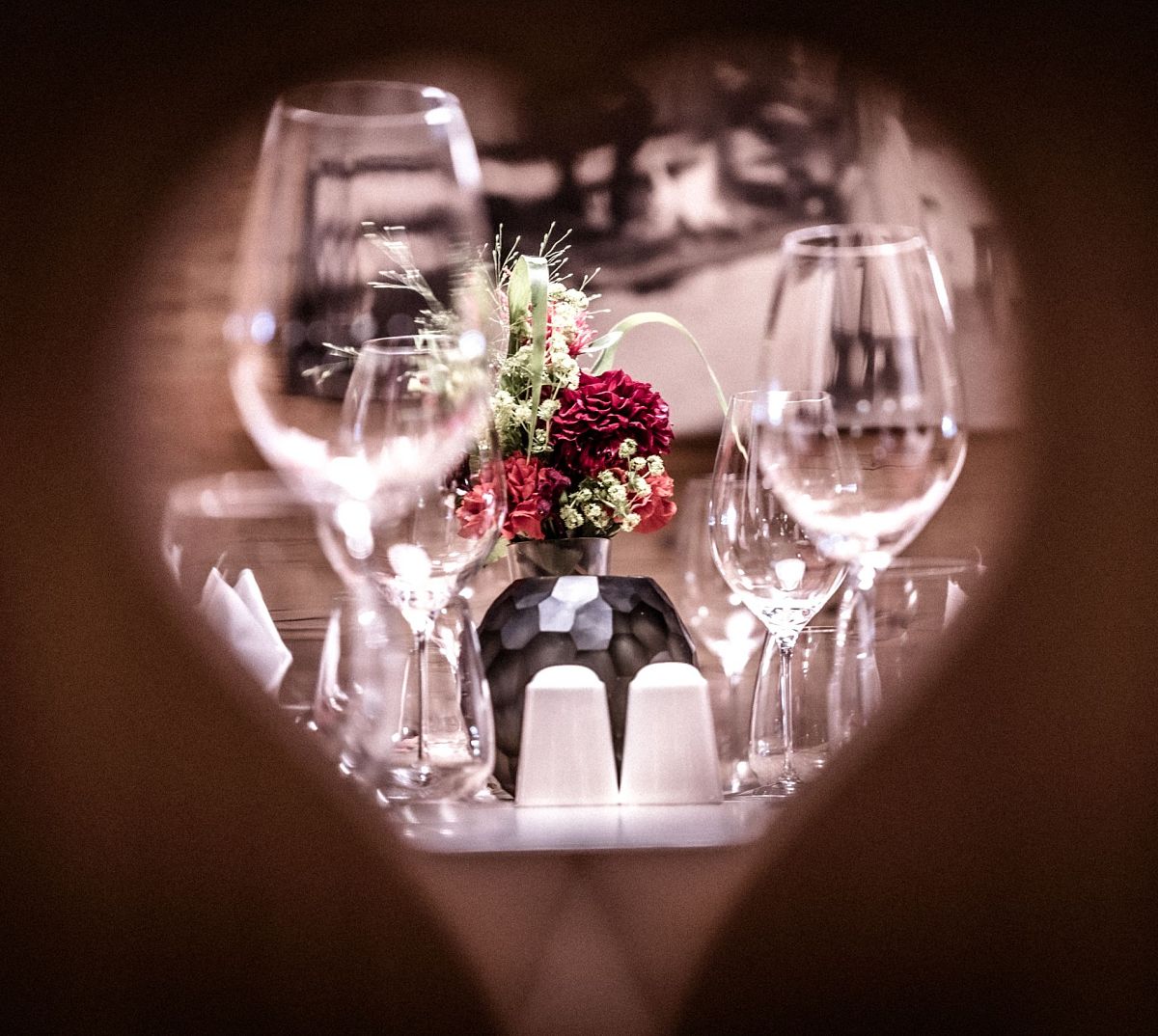 Ein Herzrahmen zeigt eine elegante Tischdekoration mit Weingläsern, einer Vase mit roten und weißen Blumen sowie Salz- und Pfefferstreuern.