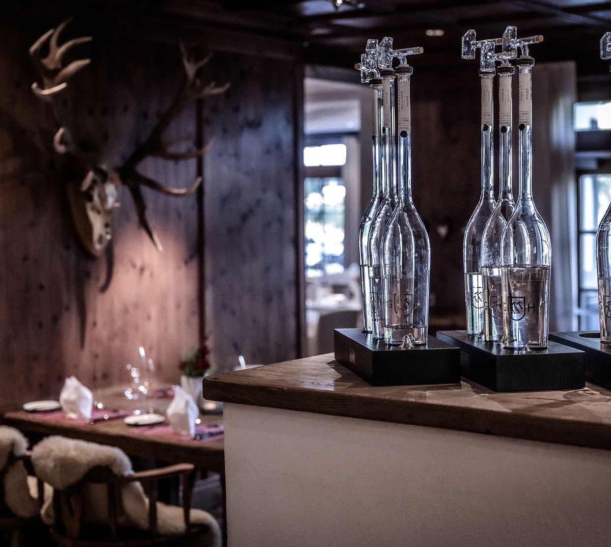 Ein gemütlicher Raum mit rustikalem Holzdekor. Auf einem Tisch stehen schmale Glasflaschen. Im Hintergrund ein Tisch mit Stühlen und ein Hirschgeweih an der Wand.