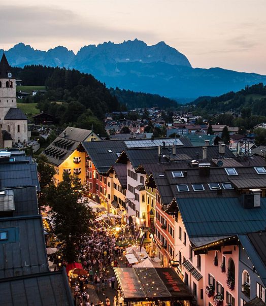 Abendstimmung in einem malerischen Bergdorf mit beleuchteten Straßen und zahlreichen Menschen. Im Hintergrund erheben sich majestätische Berge unter einem sanften Himmel.