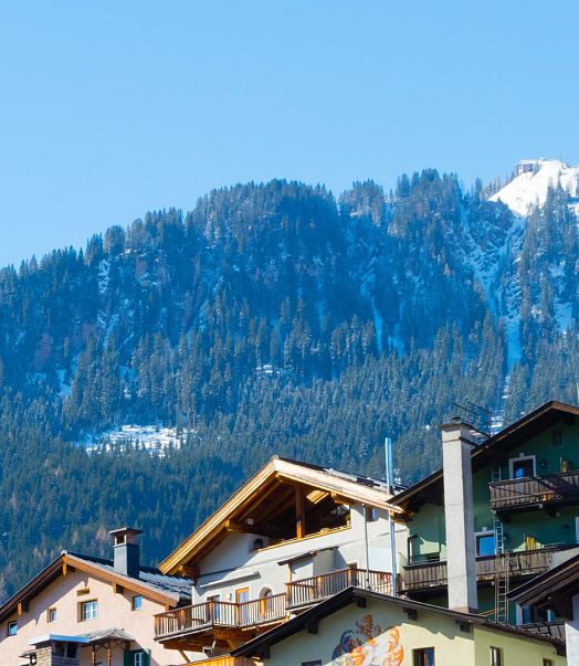 Berglandschaft mit schneebedecktem Gipfel im Hintergrund und traditionell alpinen Häusern im Vordergrund. Ein Kirchenturm ragt über die Dächer empor.