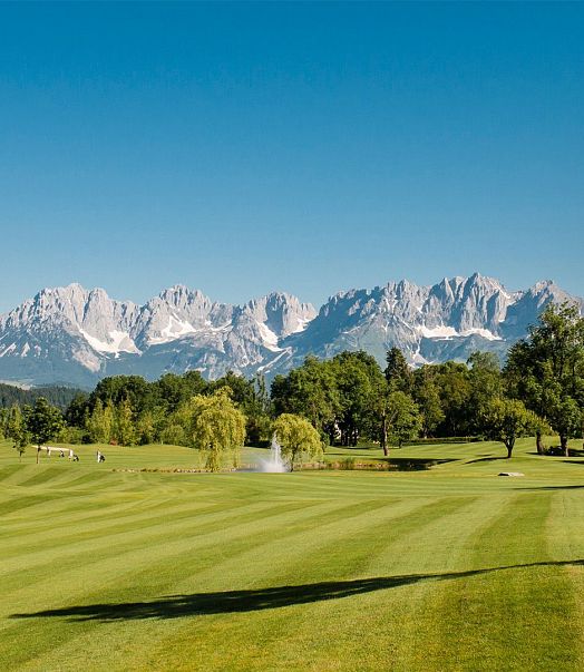 Grüne, gepflegte Golfanlage mit welligem Rasen, umgeben von üppigen Bäumen. Im Hintergrund erheben sich majestätische, schneebedeckte Alpen unter blauem Himmel.