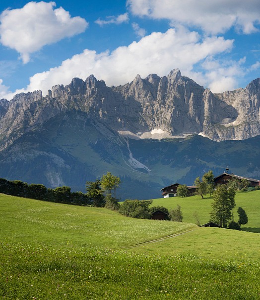 Grüne Wiese mit verstreuten Bauernhäusern im Vordergrund, imposante Bergkette unter blauem Himmel mit weißen Wolken im Hintergrund. Sommerliche, idyllische Landschaft.