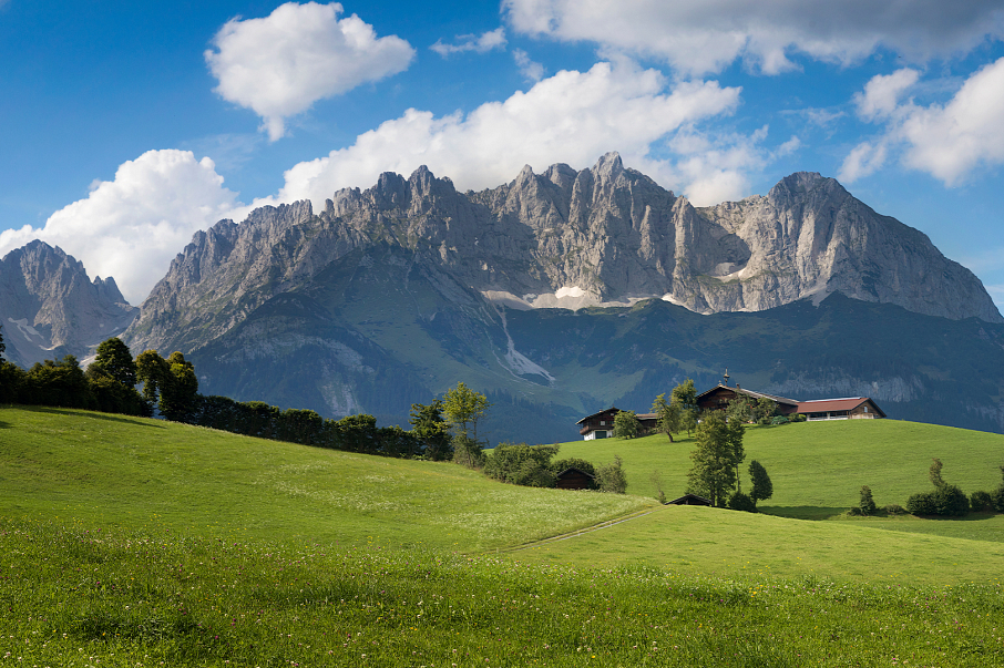 Grüne Wiese mit verstreuten Bauernhäusern im Vordergrund, imposante Bergkette unter blauem Himmel mit weißen Wolken im Hintergrund. Sommerliche, idyllische Landschaft.