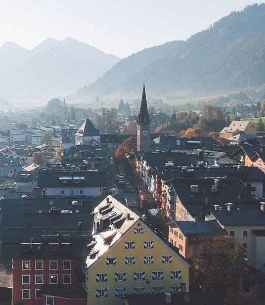 Luftaufnahme einer malerischen Stadt im Tal, umgeben von Bergen. Im Zentrum erhebt sich eine Kirche mit einem hohen Turm, umgeben von traditionellen Häusern.