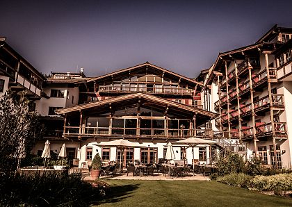 Ein großes, rustikales Hotel im Alpenstil mit Holzelementen und Balkonen, eingebettet in eine grüne Landschaft unter einem klaren, blauen Himmel.