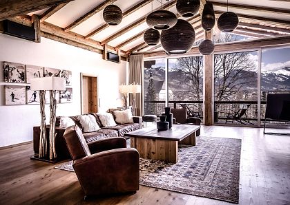 Modernes Wohnzimmer mit Holzbalkendecke und großen Fenstern, die einen Blick auf schneebedeckte Berge bieten. Gemütliche Ledermöbel und stilvolle Hängelampen.