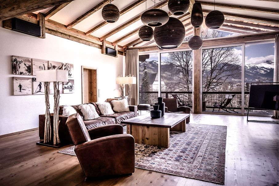 Modernes Wohnzimmer mit Holzbalkendecke und großen Fenstern, die einen Blick auf schneebedeckte Berge bieten. Gemütliche Ledermöbel und stilvolle Hängelampen.