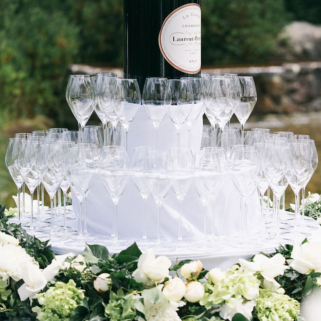 Ein eleganter Turm aus leeren Champagnergläsern auf einem Tisch, geschmückt mit weißen Blumen und Grün. Im Hintergrund ein Teich in einem Garten.