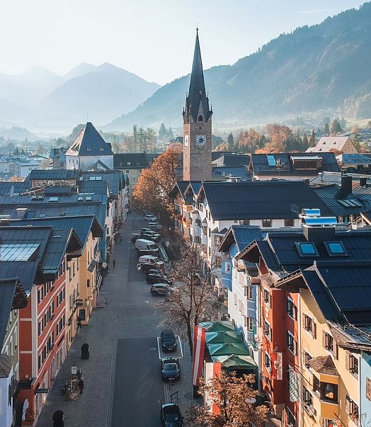 Luftaufnahme einer malerischen Stadt mit bunten Häusern und einer zentralen Kirche mit Turm. Die umliegenden Berge sind im Hintergrund zu sehen.