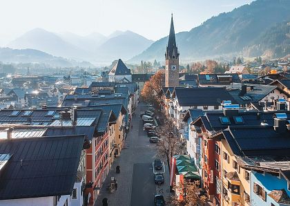 Luftaufnahme einer malerischen Stadt mit bunten Häusern und einer zentralen Kirche mit Turm. Die umliegenden Berge sind im Hintergrund zu sehen.