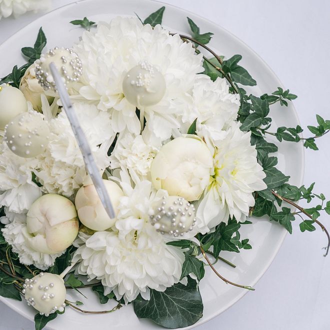 Ein eleganter Blumenstrauß mit weißen Blumen und grünen Blättern auf einem weißen Teller, auf einem weiß gedeckten Tisch mit Holzkanten angerichtet.