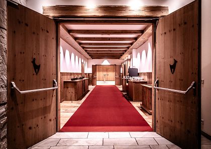 Ein eleganter Eingangsbereich mit zwei offenen Holztüren, die zu einem roten Teppich führen. Die Wände sind weiß, und Deckenstrahler sorgen für warmes Licht.