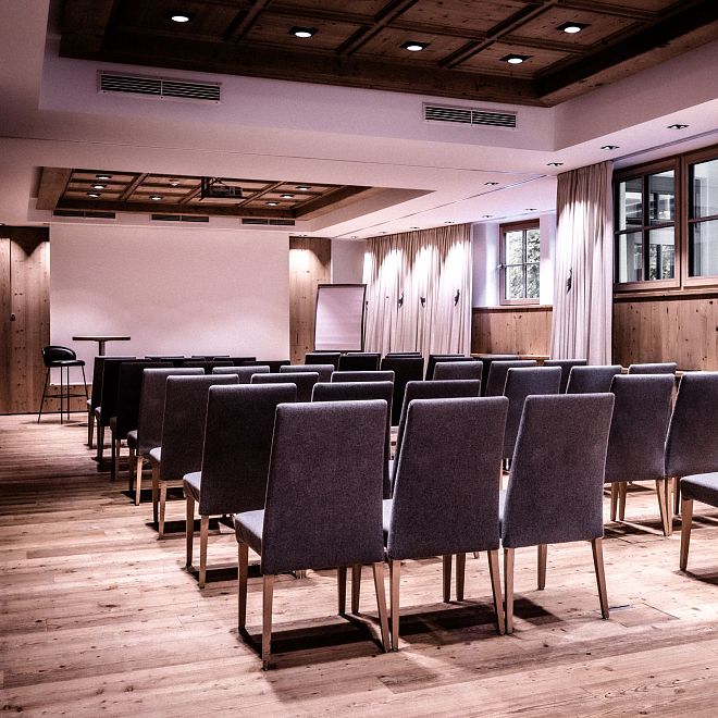 Ein moderner Konferenzraum mit Holzfußboden, Holzwänden und grauen Stühlen, die in Reihen aufgestellt sind. Ein Rednerpult und eine Leinwand sind sichtbar.