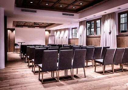 Ein moderner Konferenzraum mit Holzfußboden, Holzwänden und grauen Stühlen, die in Reihen aufgestellt sind. Ein Rednerpult und eine Leinwand sind sichtbar.